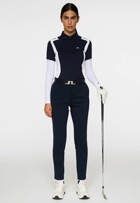 Mörkblå pikétröja med vita detaljer och långa ärmar, kombinerad med mörkblå byxor och ett svart skärp. En golfklubba i handen, vita handskar framhäver stilen.