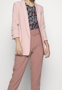 Žena v světle růžovém blejzru, květinové blůze a kalhotách s vysokým pasem v jemné růžové barvě, doplněných odpovídajícím páskem.