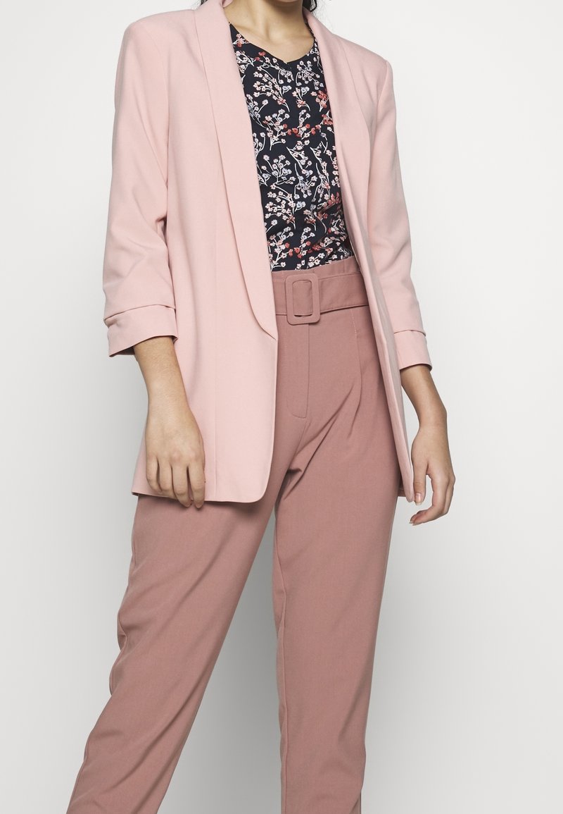 Žena v světle růžovém blejzru, květinové blůze a kalhotách s vysokým pasem v jemné růžové barvě, doplněných odpovídajícím páskem.