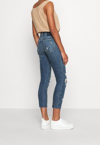 Jeans skinny blu in denim con dettagli strappati, vita alta e orlo grezzo. Abbinati a un top senza maniche beige e sneakers bianche.