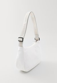 Bolso de hombro de tela blanca con forma curvada, cierre de cremallera y una correa gris claro que presenta "TOMMY JEANS" en beige.