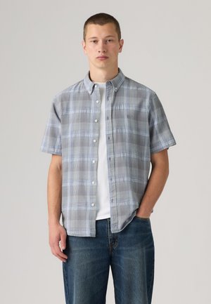 Jeune homme portant une chemise à carreaux grise à manches courtes boutonnée sur un t-shirt blanc avec un jean bleu, debout devant un fond uni.