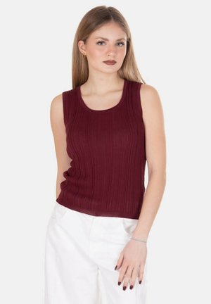 Giovane donna con capelli biondi lisci indossa un top bordeaux senza maniche a coste e pantaloni bianchi, posando su uno sfondo chiaro e uniforme.