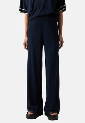 Person iført løstsiddende mørkeblå bukser, matchende top med hvid ærmekant og sorte platformsandaler mod en ensfarvet baggrund.