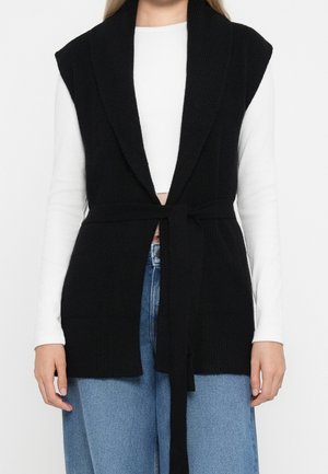 Femme portant un gilet en maille noir sans manches avec une ceinture par-dessus un haut blanc à manches longues et un jean bleu, montrée des épaules jusqu'aux hanches.