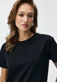 Czarna koszulka z okrągłym dekoltem, ozdobiona rozrzuconymi, małymi metalicznymi ćwiekami na przodzie. Krótkie rękawy i miękka faktura materiału.
