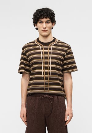 Jeune homme aux cheveux bouclés portant une chemise à manches courtes rayée marron et un pantalon marron foncé texturé avec cordon de serrage, debout devant un fond uni.