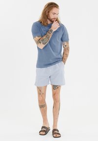 Blå bomullströja med tryckt logotyp, i kombination med ljusrandiga badshorts. Bruna sandaler fulländar den avslappnade sommaroutfiten.