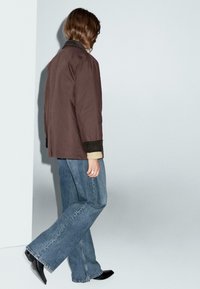 Giacca marrone con design a colletto, abbinata a jeans a gamba larga azzurri. La giacca presenta polsini scuri e un tessuto testurizzato. Scarpe nere a punta.