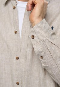 Beige gestreifte Hemdbluse mit braunen Knöpfen, ausgestattet mit einem taillierten Kragen und langen Ärmeln. Texturierter Stoff mit subtilen Musterdetails.