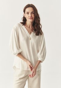 Bluză crem cu decolteu în V, mâneci bufante gathered, textură subtilă și tiv rotunjit; asociată cu pantaloni bej deschis.