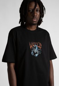 Melna kokvilnas T-krekls ar krāsainu skorpiona grafiku un vārdu "WASTED" virsū oranžā krāsā. Īsas piedurknes, brīvs piegriezums.