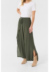 Gonna maxi verde oliva con vita regolabile, spacchi laterali, realizzata in tessuto leggero; abbinata a una blusa bianca con bordi ondulati e stivaletti beige alla caviglia.