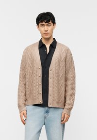 Νέος άνδρας που φορά ένα μπεζ πουλόβερ πλέξης καλωδίου πάνω από μαύρο πουκάμισο και ανοιχτό μπλε τζιν, στέκεται μπροστά σε ένα απλό λευκό φόντο.