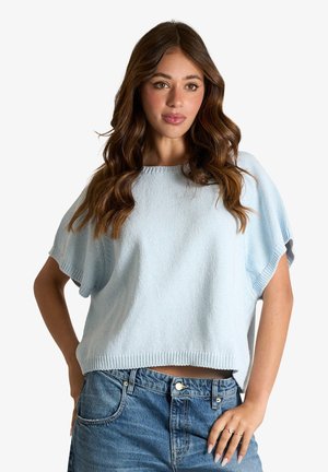 Donna con lunghi capelli mossi castani che indossa un maglione azzurro a maniche corte e jeans blu, posando con una mano sul fianco e sfondo neutro.