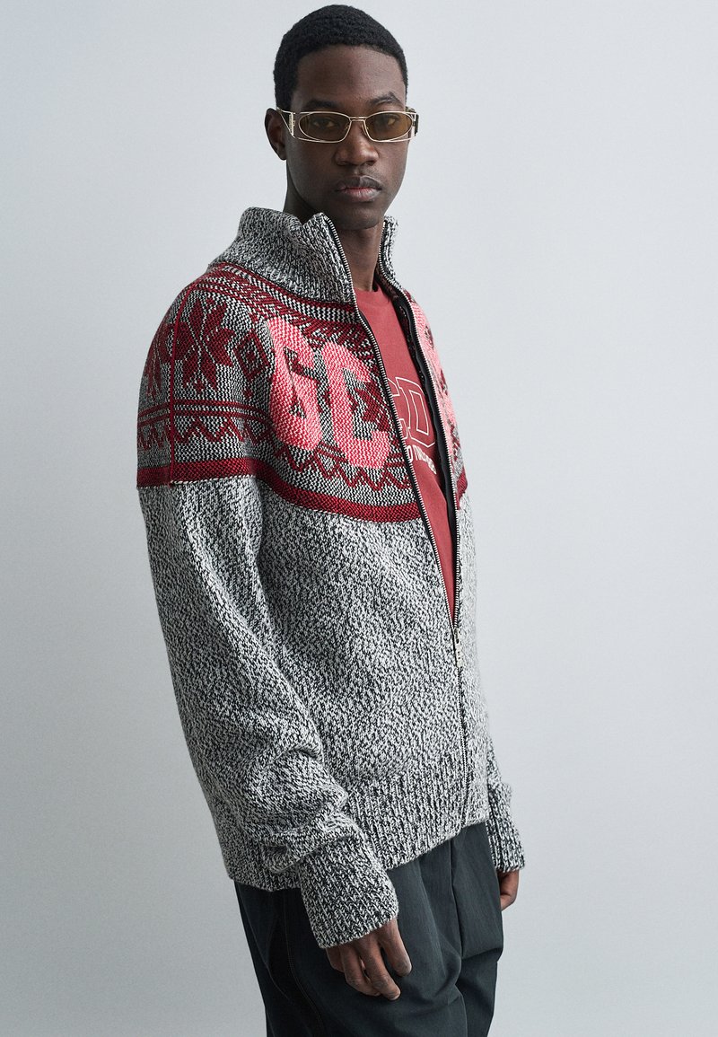 Grijze gebreide ritssweater met een rode gebreide yoke. Heeft dikke manchetten en een hoge kraag. Gekozen boven een rood T-shirt.