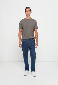 En ung mann står mot en hvit bakgrunn iført grå t-skjorte med korte ermer, blå jeans og hvite joggesko.