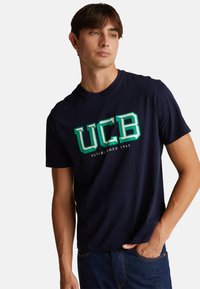 Námořnická modrá tričko UCB vyrobené z bavlny, s zeleným logem "UCB" a textem "ESTABLISHED 1965", krátkými rukávy a kulatým výstřihem.