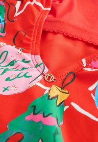 Czerwona tkanina z kolorowymi, zabawnymi wzorami choinek i ozdób świątecznych. Dekolt w kształcie litery V z złotym akcentem przy linii szyi.