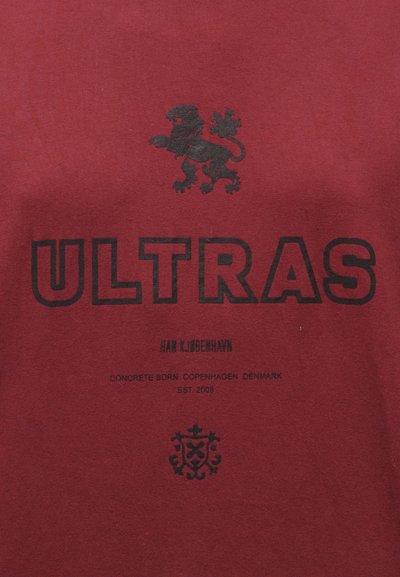 T-shirt vermelha de algodão com gráfico de leão preto, texto em destaque "ULTRAS" e texto adicional em preto, com uma textura suave subtil.
