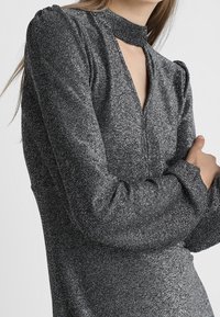 Glittrig svart topp med en hög choker-krage, långa ärmar och en nyckelhål-detail vid halslinningen. Mjuk, texturerad tyg med en skimrande effekt.