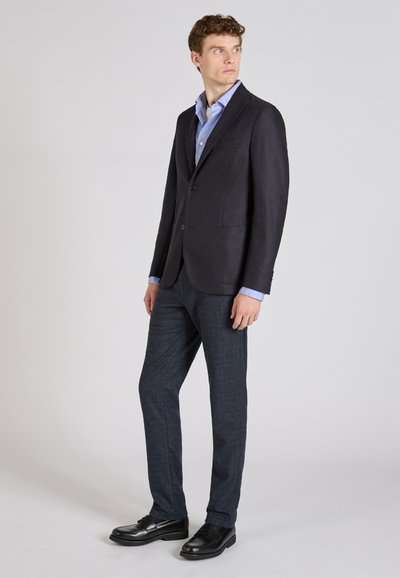 Dopasowana ciemna marynarka na jasnoniebieską koszulę, zestawiona z granatowymi spodniami i czarnymi mokasynami. Marynarka ma dwa przednie kieszenie i klapy w kształcie notcha.