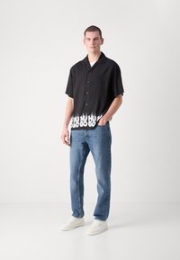 Cămașă neagră cu mânecă scurtă, cu un grafic alb în flăcări la tiv, asociată cu blugi albaștri și teniși albi. Material neted, stil de guler clasic.
