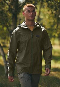 Veste imperméable vert olive avec capuche, fermeture éclair à l'avant et poignets ajustables, portée sur un haut jaune. Matériau léger, cadre extérieur.
