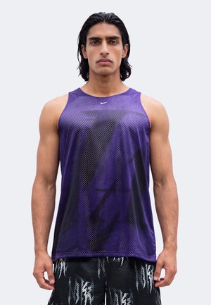 Joven con pelo oscuro lleva una camiseta deportiva morada sin mangas y pantalones cortos negros con estampado sobre un fondo claro y liso.