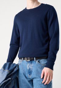 Maglione di maglia blu navy con maniche lunghe, collo rotondo e discreto logo. Abbinato a jeans di denim azzurro e con in mano una giacca di denim.