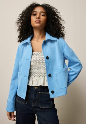 Woman with curly hair wearing light blue buttoned jacket over white textured top and dark blue jeans, standing against beige wall.