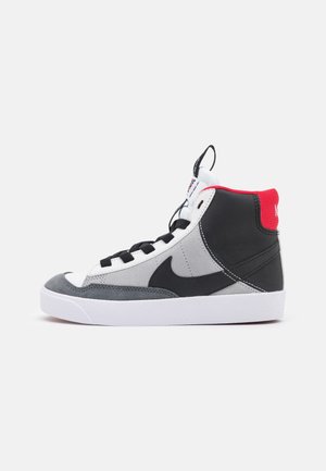 Hoher Sneaker mit schwarzen, weißen und grauen Paneelen, schwarzen Schnürsenkeln, rotem Fersenkragen und weißer Sohle, seitlich auf einem einfarbigen Hintergrund gezeigt.