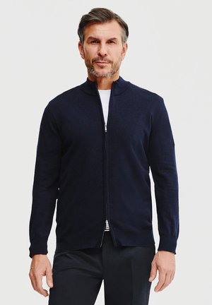Granatowy sweter zapinany na zamek z miękkiego materiału dżersejowego, z wysokim kołnierzem, dopasowany krój i subtelny połysk. Posiada zamek z przodu i minimalne detale.