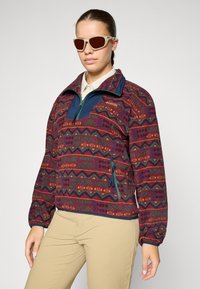 Pullover in felpa bordeaux con motivi geometrici in verde, blu e giallo. Presenta un colletto blu e una tasca con zip sul davanti.