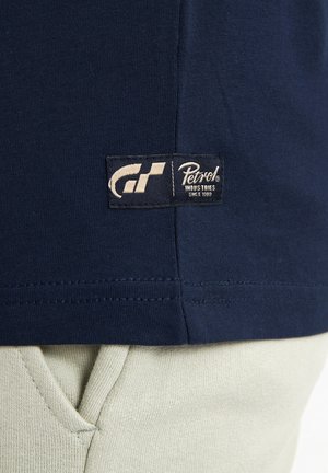T-shirt in cotone blu navy con etichetta visibile che presenta "Petrol" e un logo da corsa. La texture è liscia con una vestibilità regolare.
