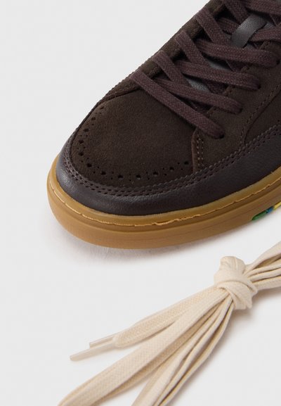 Κοντινό πλάνο στην μύτη ενός καφέ σουέντ και δερμάτινου αθλητικού παπουτσιού με σκουρόχρωνα καφέ κορδόνια και δίπλα ένα ζευγάρι μπεζ κολλημένων κορδονιών σε λευκό φόντο.