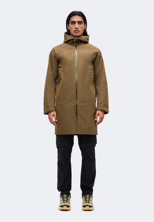 Mann steht und trägt eine olivgrüne lange Kapuzenjacke, schwarze Hose und beige-schwarze Wanderschuhe vor einem einfarbigen hellen Hintergrund.