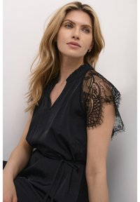Schwarzes Kleid mit kurzen Spitzensäumen, V-Ausschnitt, strukturiertem Stoff und einem Bindegürtel in der Taille. Die Spitze hat florale Muster.