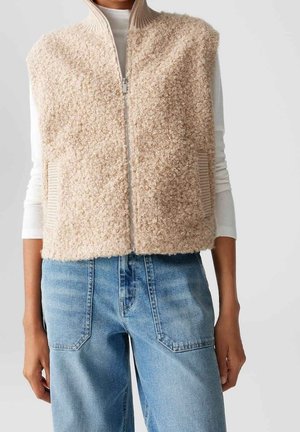 Personne portant un gilet beige texturé zippé sur une chemise blanche à manches longues et un jean bleu clair, visible du cou jusqu'aux hanches.