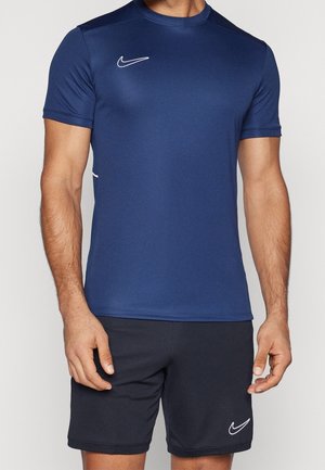 Homme portant un maillot de sport Nike bleu marine et un short Nike noir, debout avec les bras détendus le long du corps devant un fond uni.