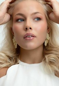 Guldstatementörhängen med en texturerad, bladliknande design. Slät, glansig hud och lätt smink. Vit topp med hög hals.