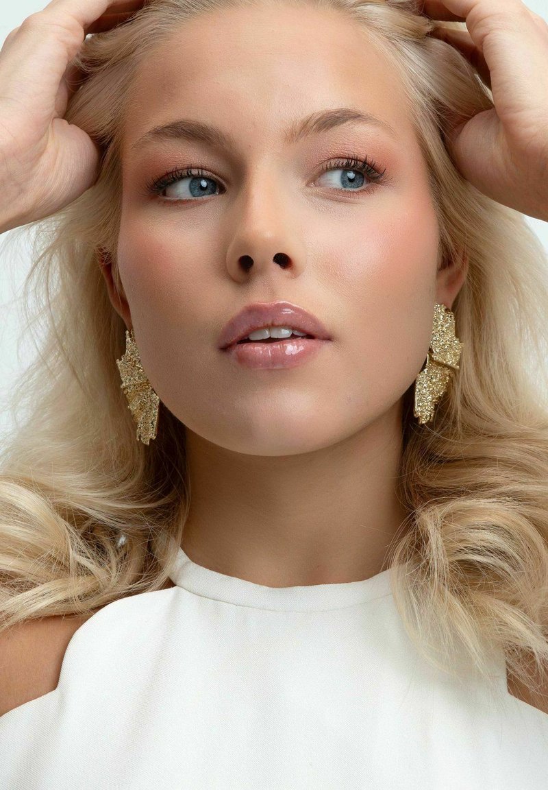 Guldstatementörhängen med en texturerad, bladliknande design. Slät, glansig hud och lätt smink. Vit topp med hög hals.