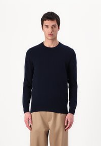 Pull bleu marine à col rond en matériau doux, avec des manches longues et des poignets côtelés, accompagné de pantalons beiges. Design simple et ajusté.