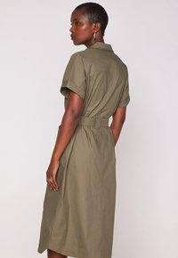 Abito camicia kaki con colletto, maniche corte, cintura in vita e tasche laterali. Tessuto liscio e textured con un taglio a lunghezza ginocchio.