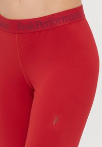Leggings atléticos vermelhos com uma faixa elástica com marca. Textura suave, design ajustado, apresentando um logo discreto na parte inferior da perna.