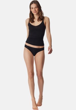 Woman with brown hair wearing a black tank top and black underwear, standing barefoot against a plain white background.