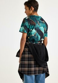 Kortärmad teal t-shirt med örntryck, kombinerad med en svart och grå rutmönstrad overshirt knuten i midjan. Avslappnade jeans.