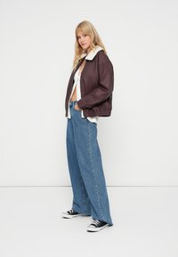 Giacca bomber in pelle marrone con colletto in shearling bianco, abbinata a jeans blu a gamba ampia e sneaker nere. Design semplice e casual.