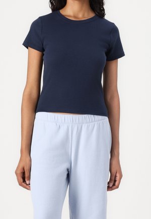 Vrouw draagt een aansluitend donkerblauw T-shirt met korte mouwen en lichtblauwe elastische taille broek staand tegen een effen achtergrond.