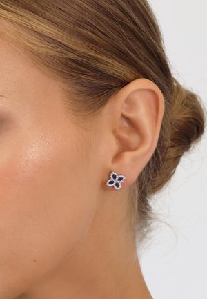 Ohr mit Ohrstecker in Form einer Blume, die vier dunkle blaue Steine zeigt, umgeben von kleinen klaren Kristallen, das Haar zu einem Dutt hochgesteckt.